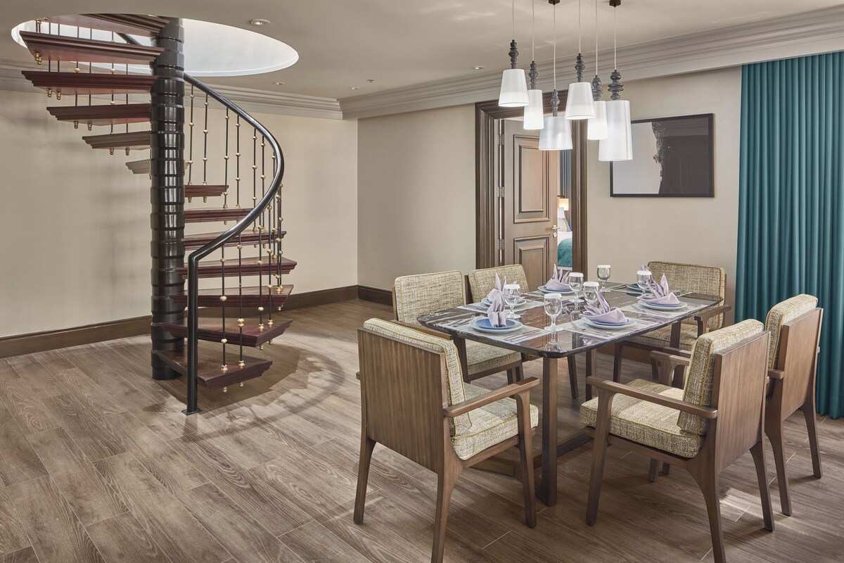 Un coin repas avec une table en bois dressée pour quatre personnes, entourée de chaises rembourrées. Un escalier en colimaçon avec des marches en bois et des rampes en métal est visible sur la gauche.