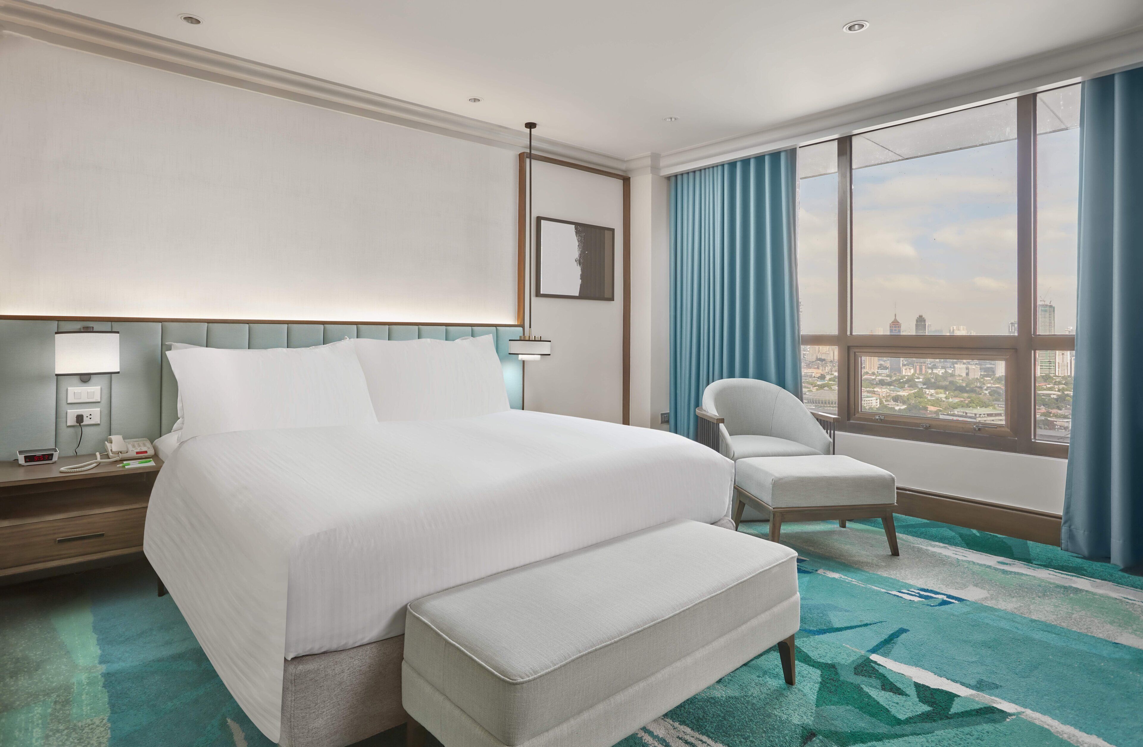 A hotel room with a large bed, a chair, a desk with a lamp, and a view of the city through large windows.