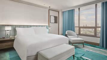 A hotel room with a large bed, a chair, a desk with a lamp, and a view of the city through large windows.