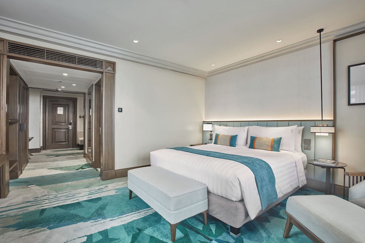 A hotel room with a large bed, a bench, a bedside table, a nightstand, a mirror, and a hallway leading to another room.