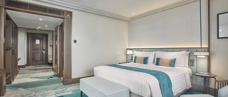 Habitación de hotel con una cama grande, un banco, una mesita de noche y un espejo, y un pasillo que conduce a otra habitación.