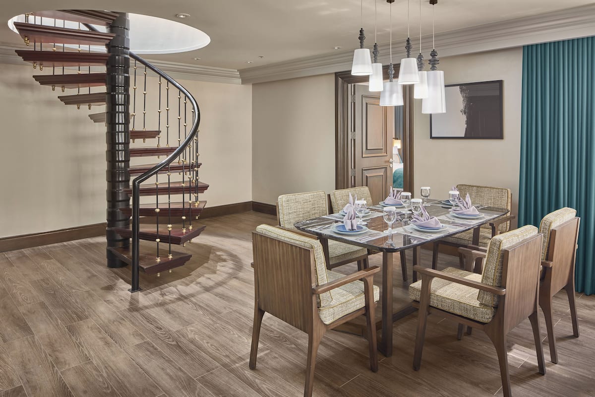 A dining area with a wooden table set for four, surrounded by upholstered chairs. A spiral staircase with wooden steps and metal railings is visible to the left.