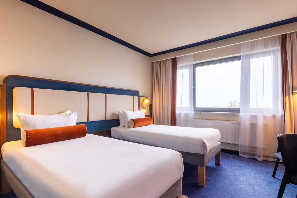 A hotel room with two beds, a chair, a window with curtains, and a radiator.
