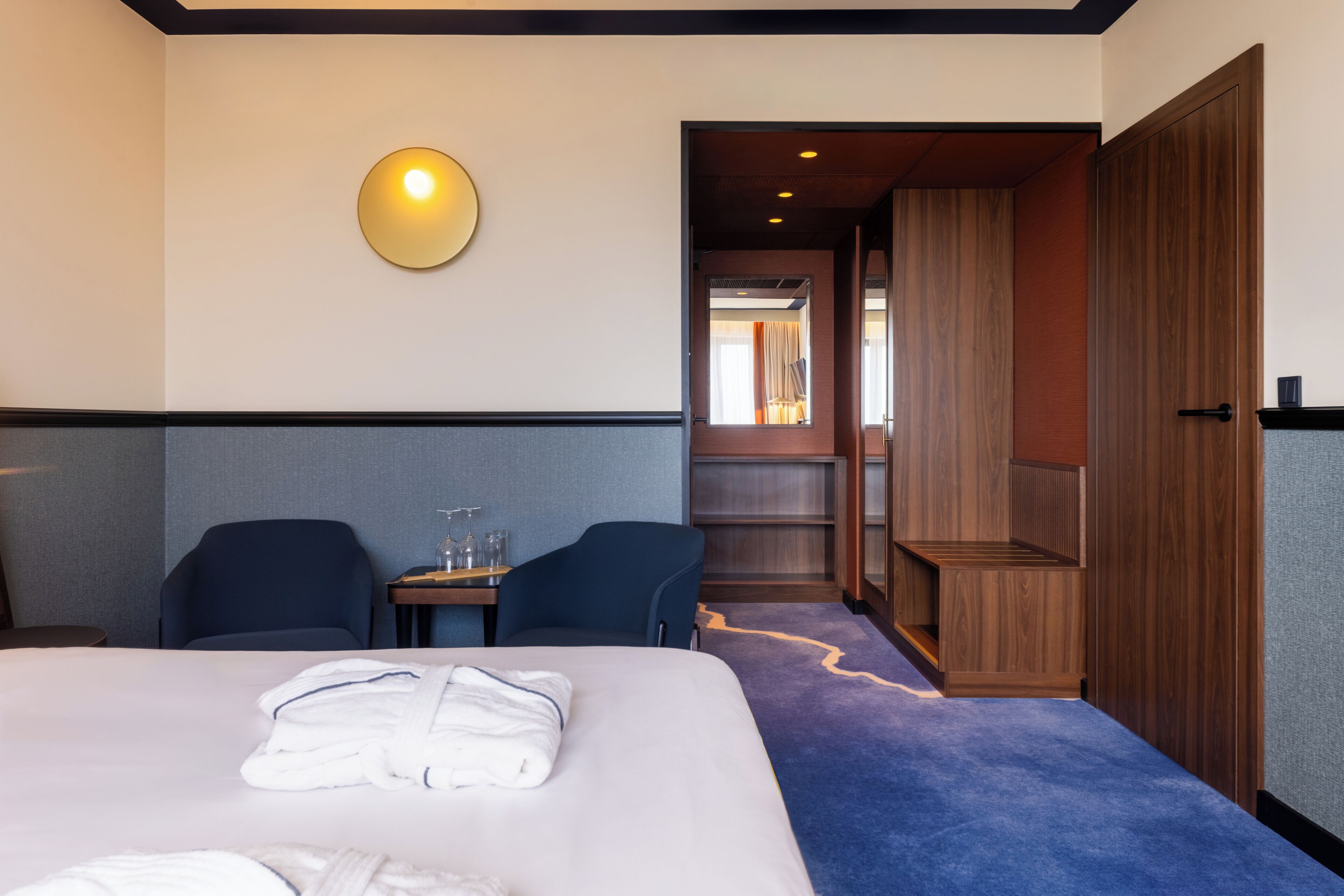 A hotel room with a bed, two chairs, a small table, a wall-mounted light, and a wooden wardrobe.