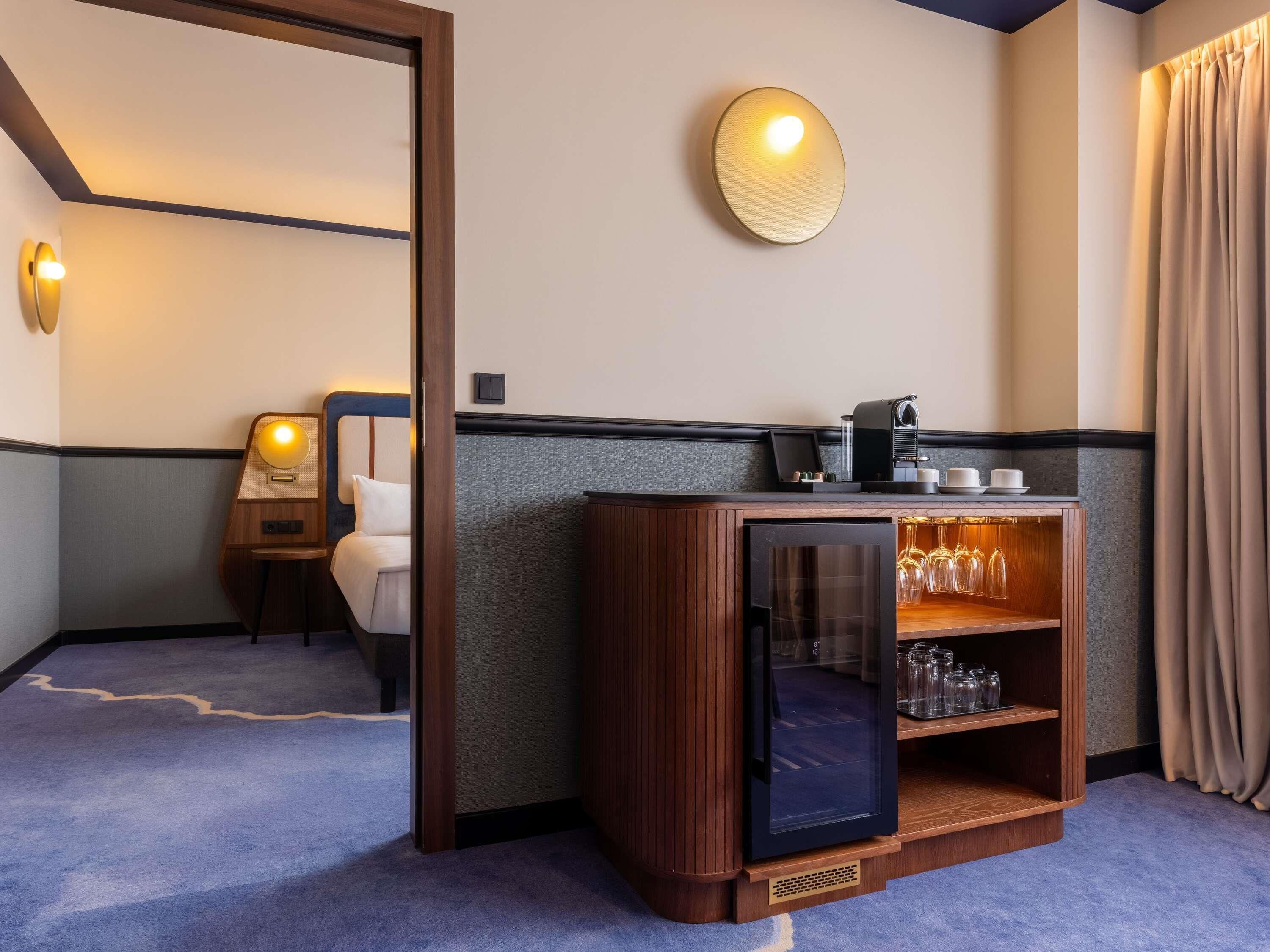 Hotellihuone, jossa on sänky, minibaari, kahvinkeitin ja lasinen vitriinikaappi.