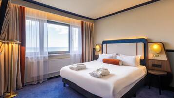 A hotel room with a large bed, two bedside lamps, a view of the sea, and a round table.