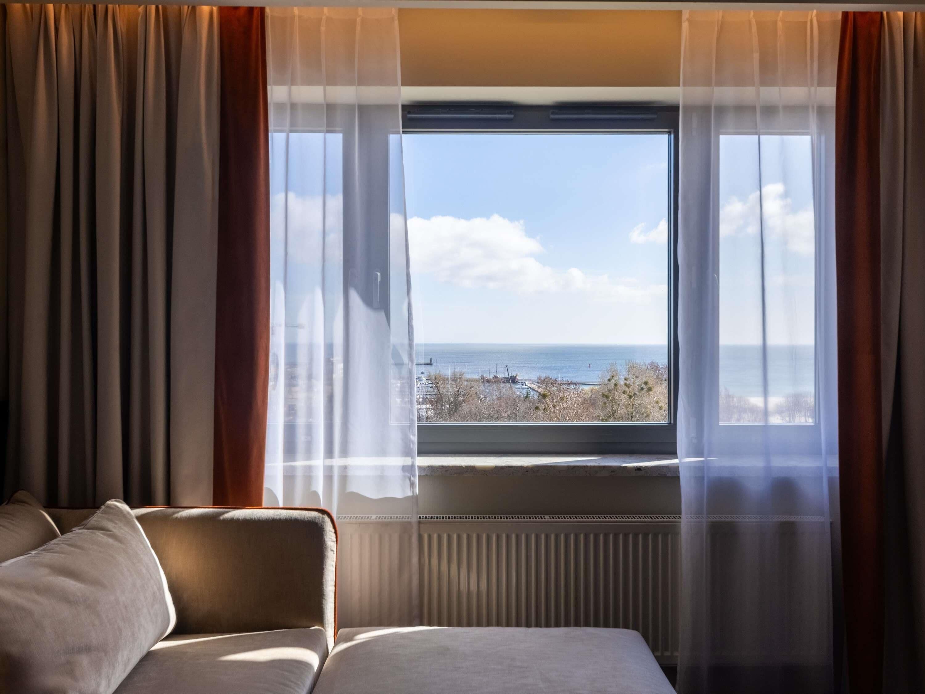 Un cuarto de hotel con una cama, un ventanal con cortinas translúcidas y vista al mar.