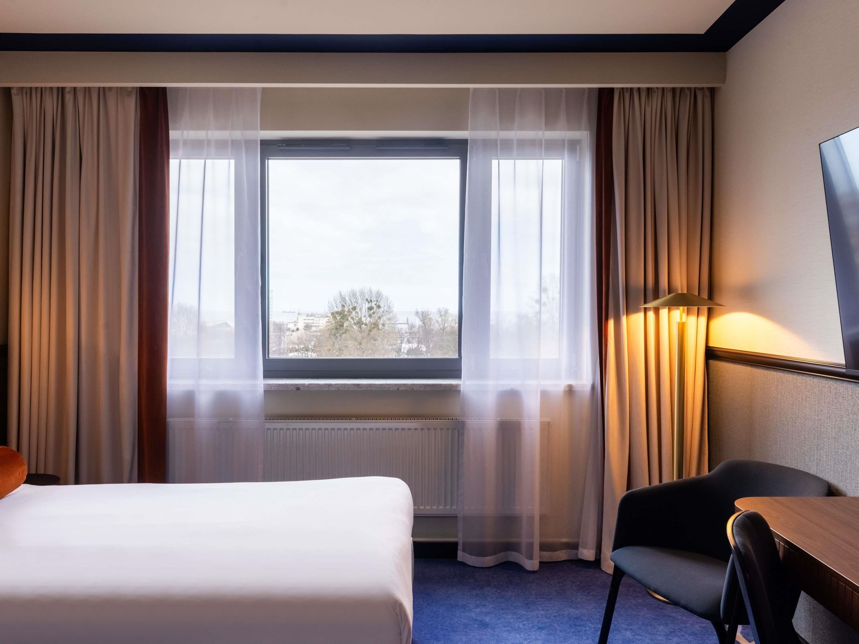 A hotel room with a bed, a chair, a table, a lamp, and a window with curtains.