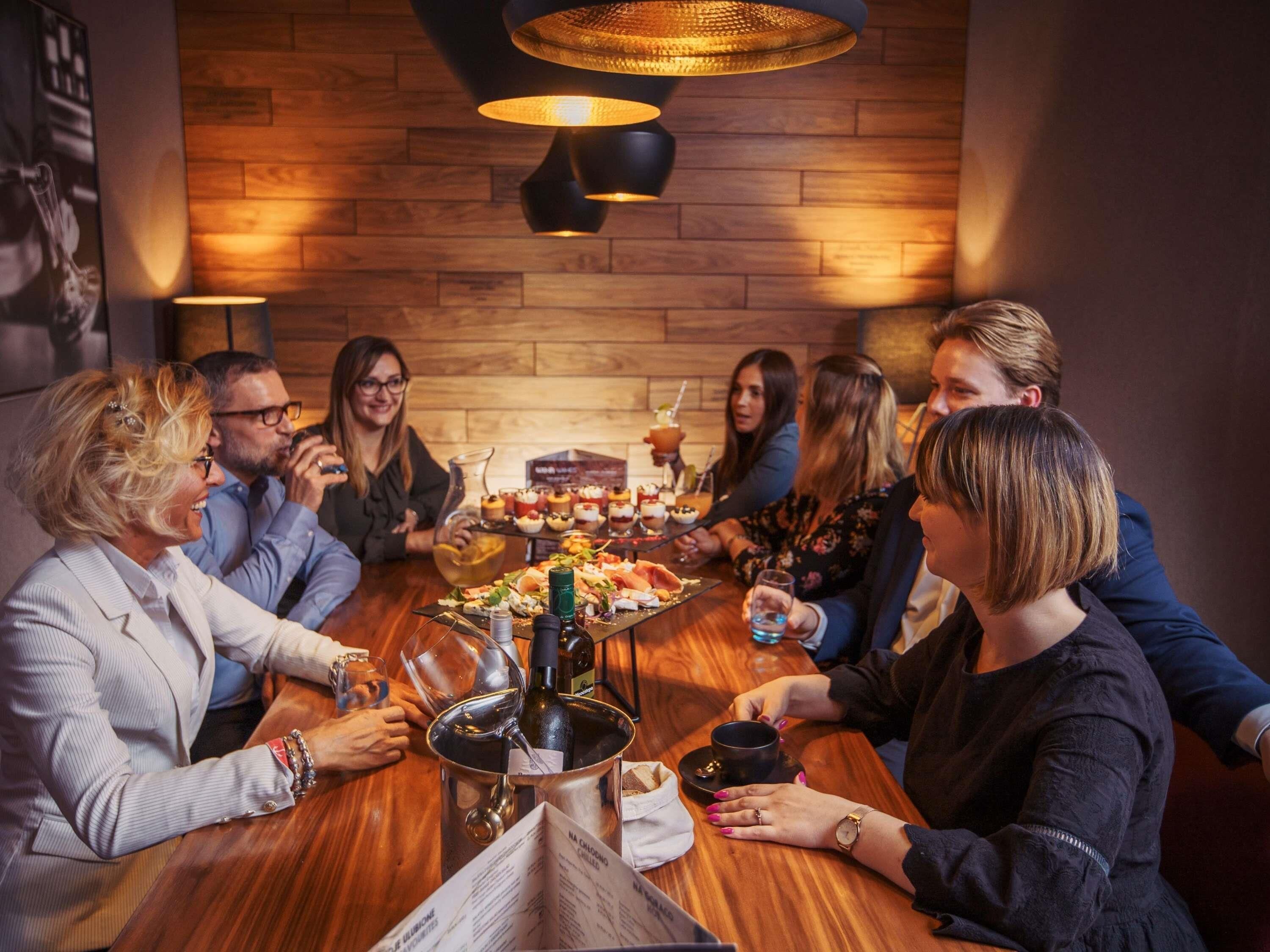 Un grupo de personas sentadas alrededor de una mesa de madera con una botella de vino, copas y un menú en una sala con paredes de madera e iluminación colgante.