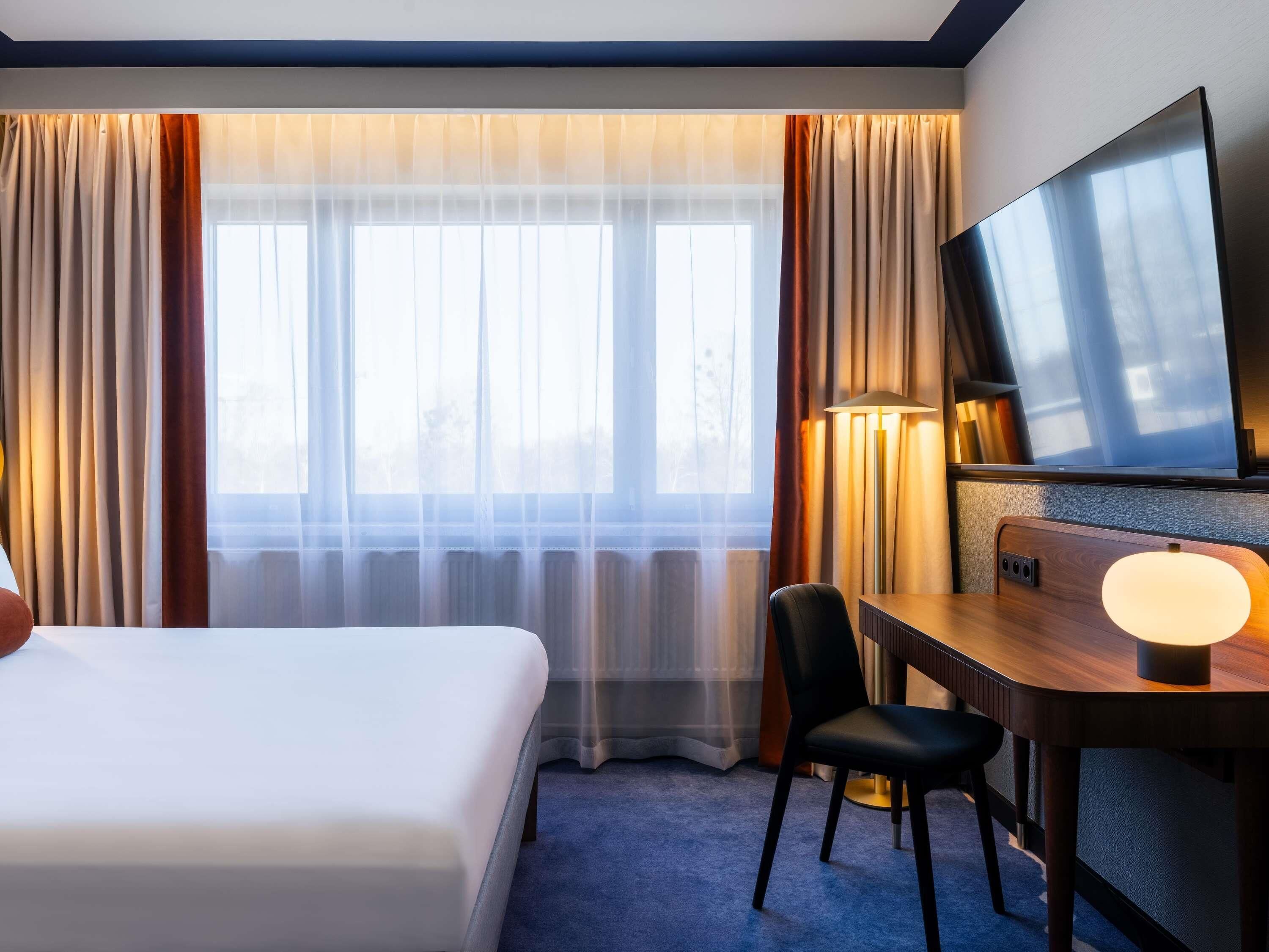 A hotel room with a bed, a desk with a chair, a television, and a lamp.