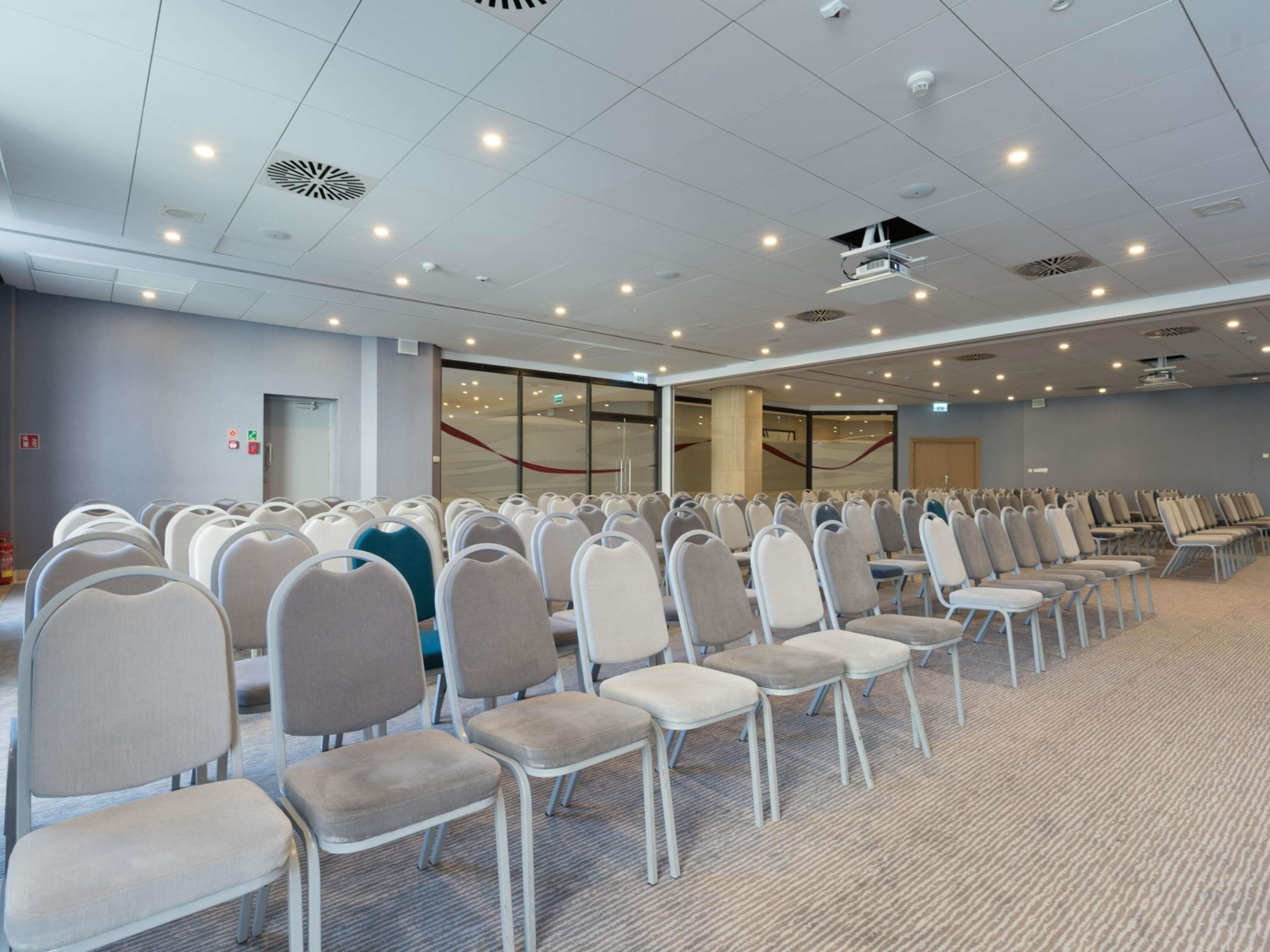 Un salón de conferencias con filas de sillas, iluminación en el techo y una pared de vidrio.