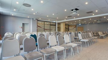 Un salón de conferencias con filas de sillas, iluminación en el techo y una pared de vidrio.