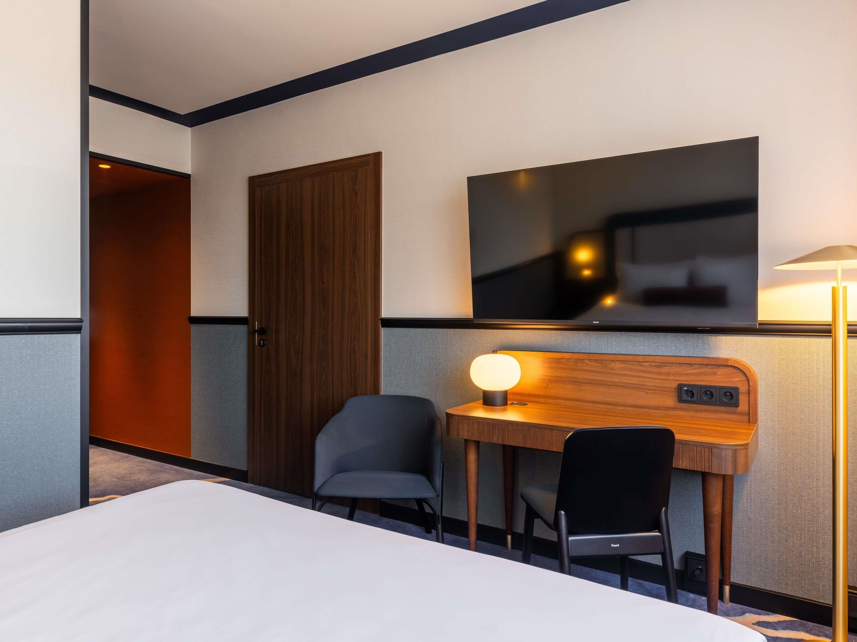 Una habitación de hotel con una cama, un escritorio con lámpara, una silla y un televisor montado en la pared.