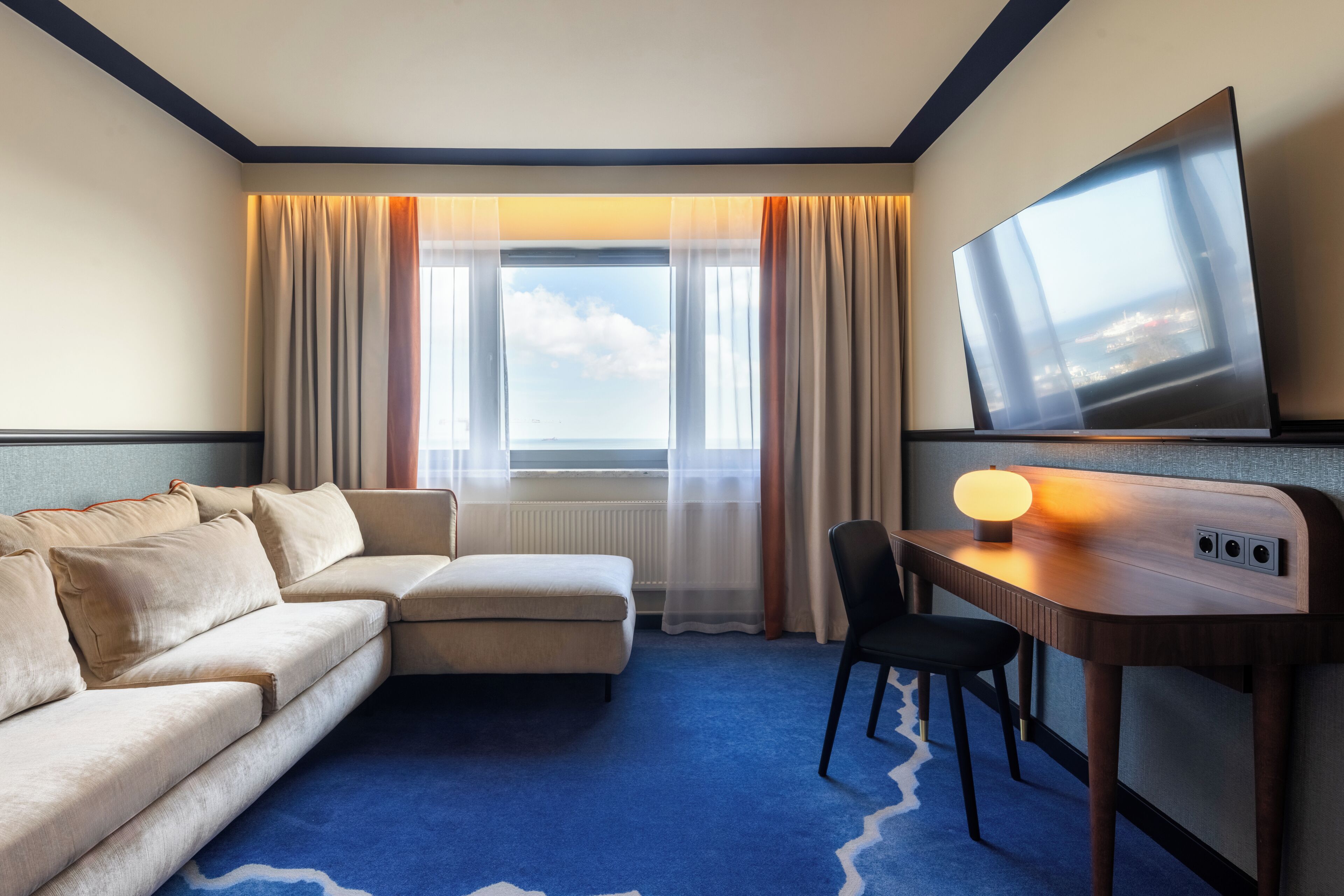 A hotel room with a sofa, a desk with a lamp, a TV, and a view of the sea through the window.