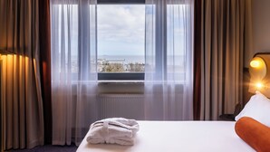 Une chambre d’hôtel avec un lit, une fenêtre avec des rideaux, une vue sur la ville et une table de chevet avec une lampe.