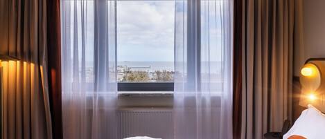A hotel room with a bed, a window with curtains, a view of the city, and a nightstand with a lamp.
