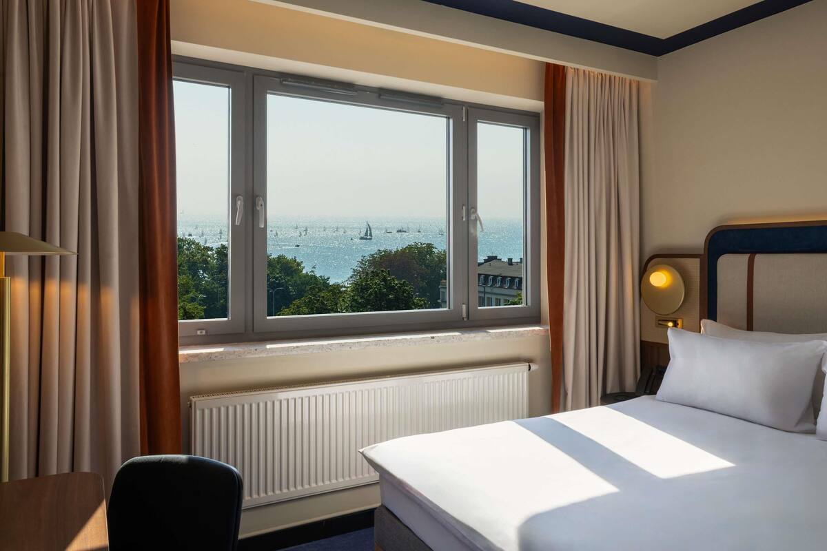 A hotel room with a large window, a bed, a chair, and a bedside table.