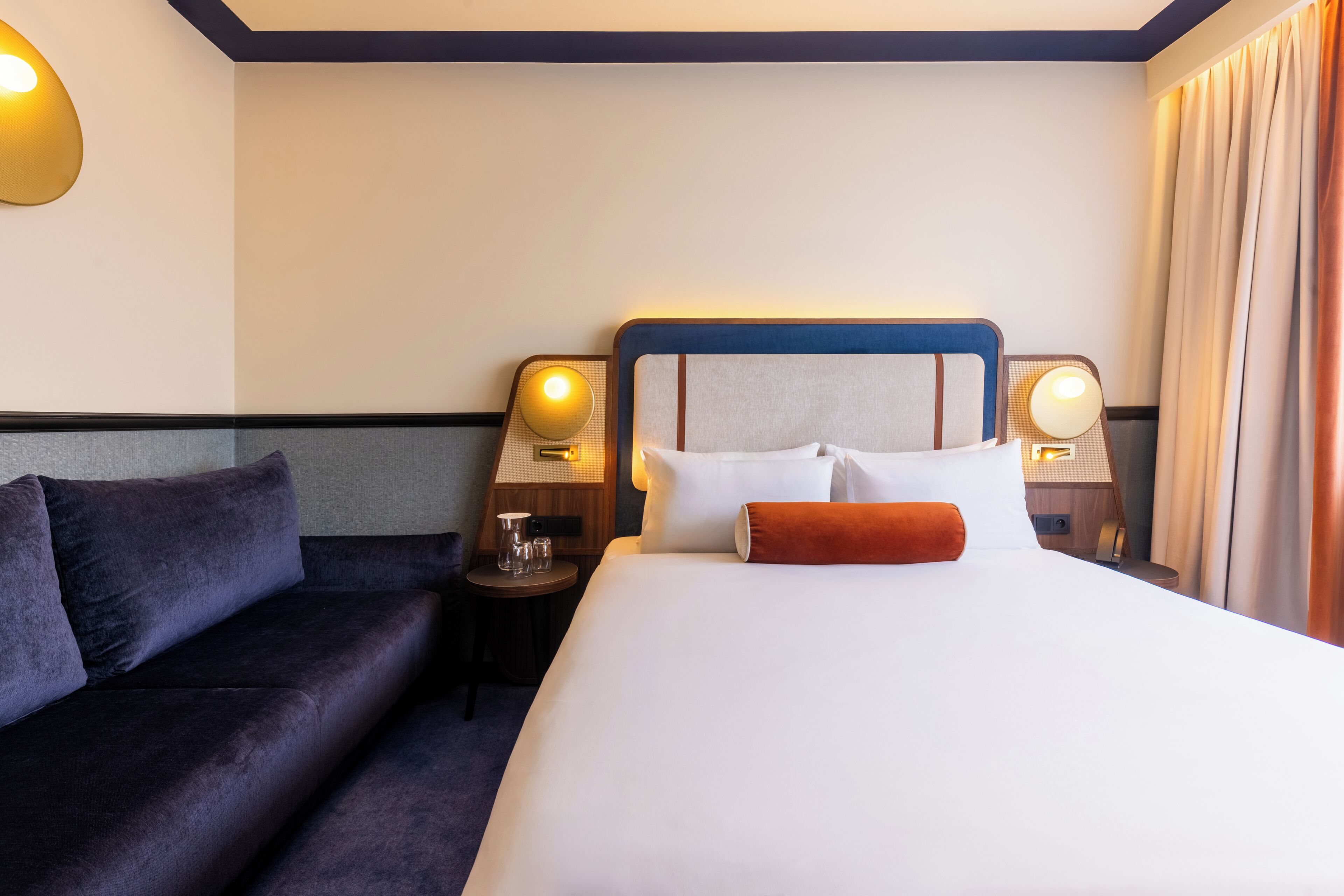 A hotel room with a bed, a sofa, two bedside lamps, and a small table with a glass.