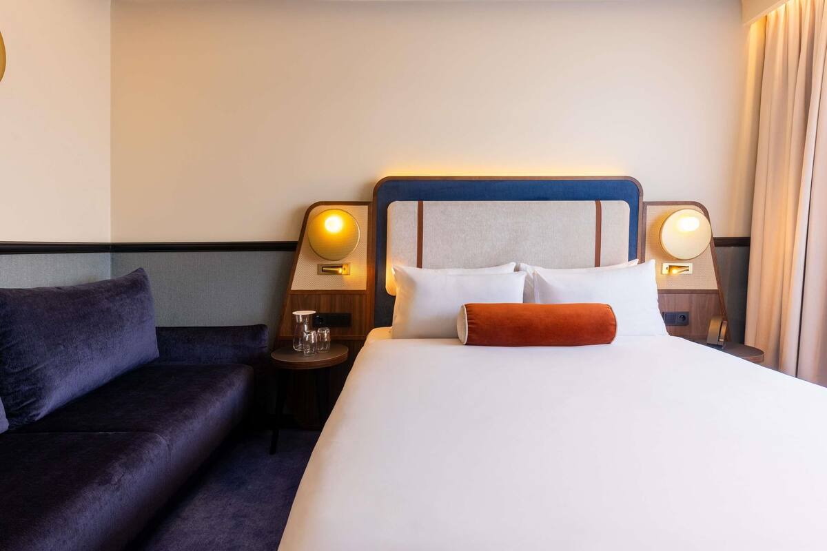 A hotel room with a bed, two bedside tables, a small round table, a sofa, and a headboard with a light fixture.