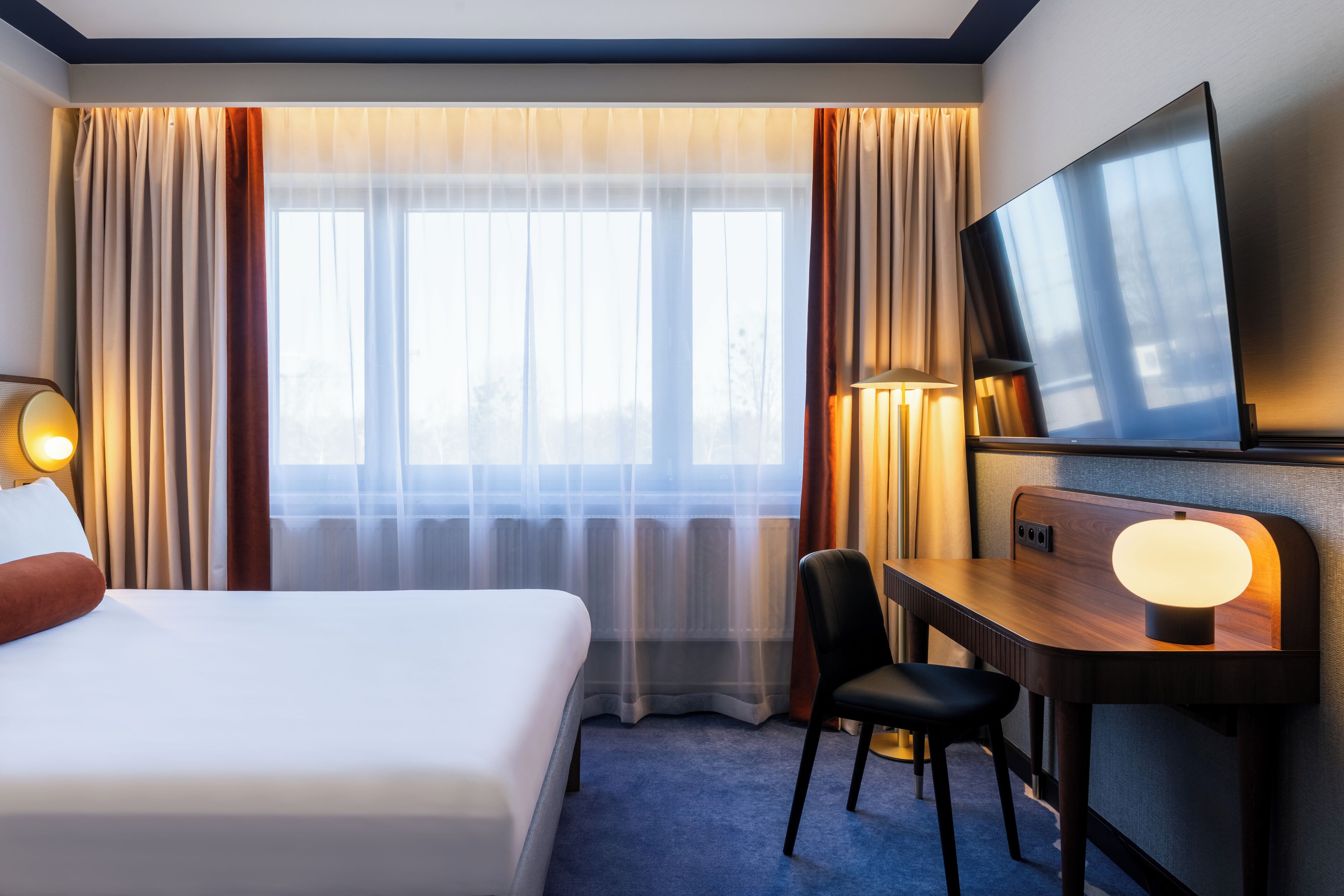 Una habitació d'hotel amb un llit, un escriptori amb una cadira, una televisió i una finestra amb cortines.