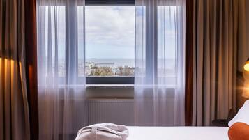 A hotel room with a bed, a window with curtains, and a view of the sea and buildings.