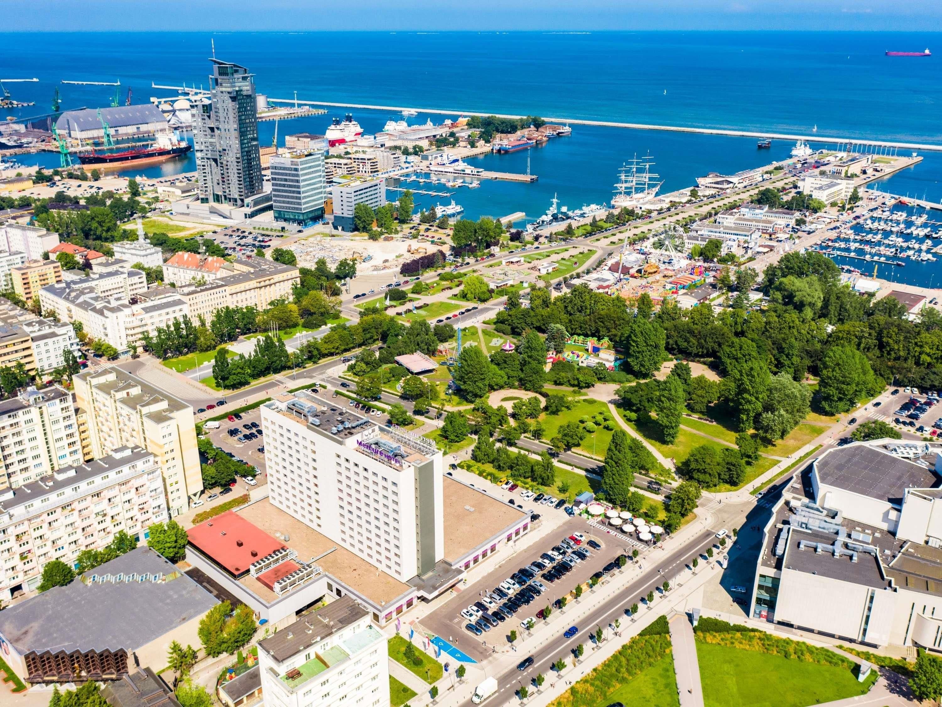Vue aérienne d’une ville côtière avec un port de plaisance, des bâtiments modernes et des espaces verts.