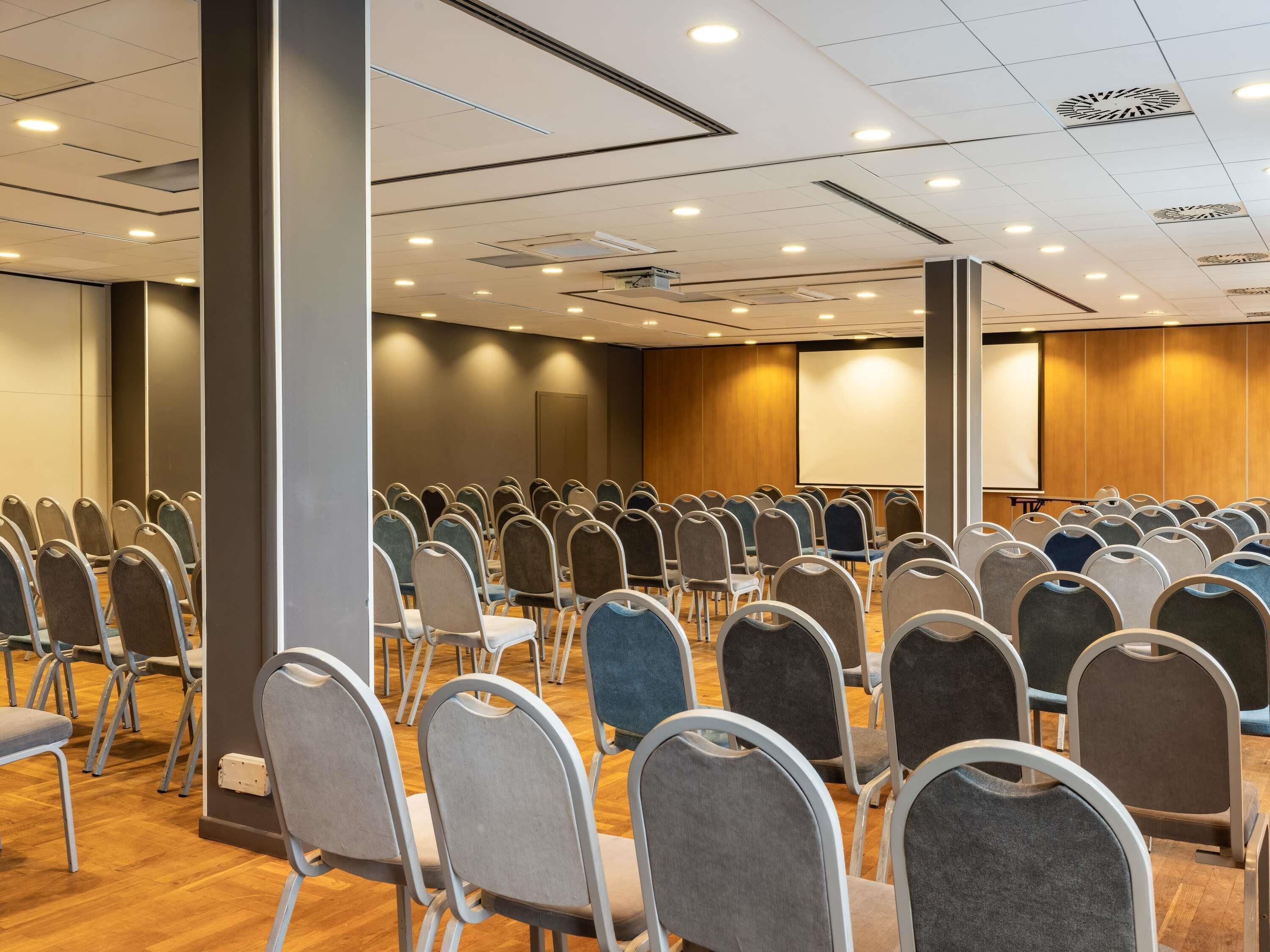 Une salle de conférence avec des rangées de chaises, un écran de projection et un mur recouvert de panneaux en bois.