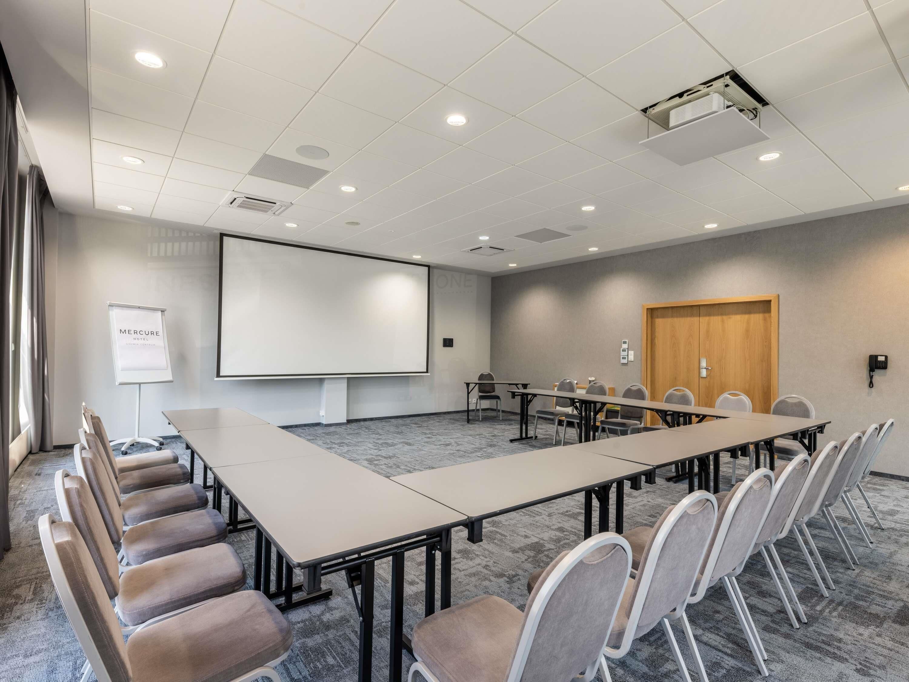 Uma sala de conferência com uma mesa comprida, cadeiras, uma tela de projetor e um quadro branco.