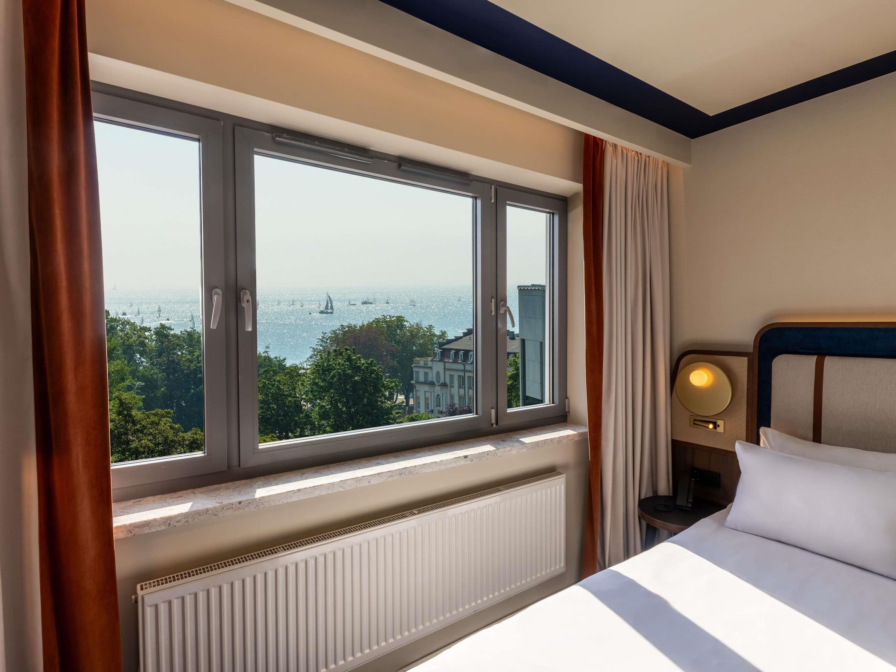 Une chambre d’hôtel avec une grande fenêtre, un lit, une table de chevet avec une lampe et un radiateur.