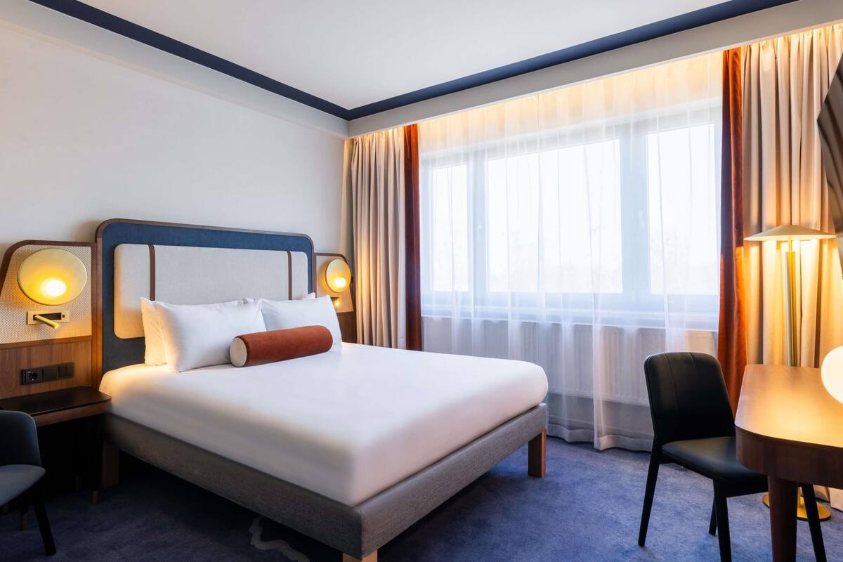 A hotel room with a large bed, two bedside tables with lamps, a desk with a chair, and a large window with curtains.