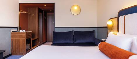 Habitación de hotel con cama, mesita de noche, lámpara y vista al pasillo.