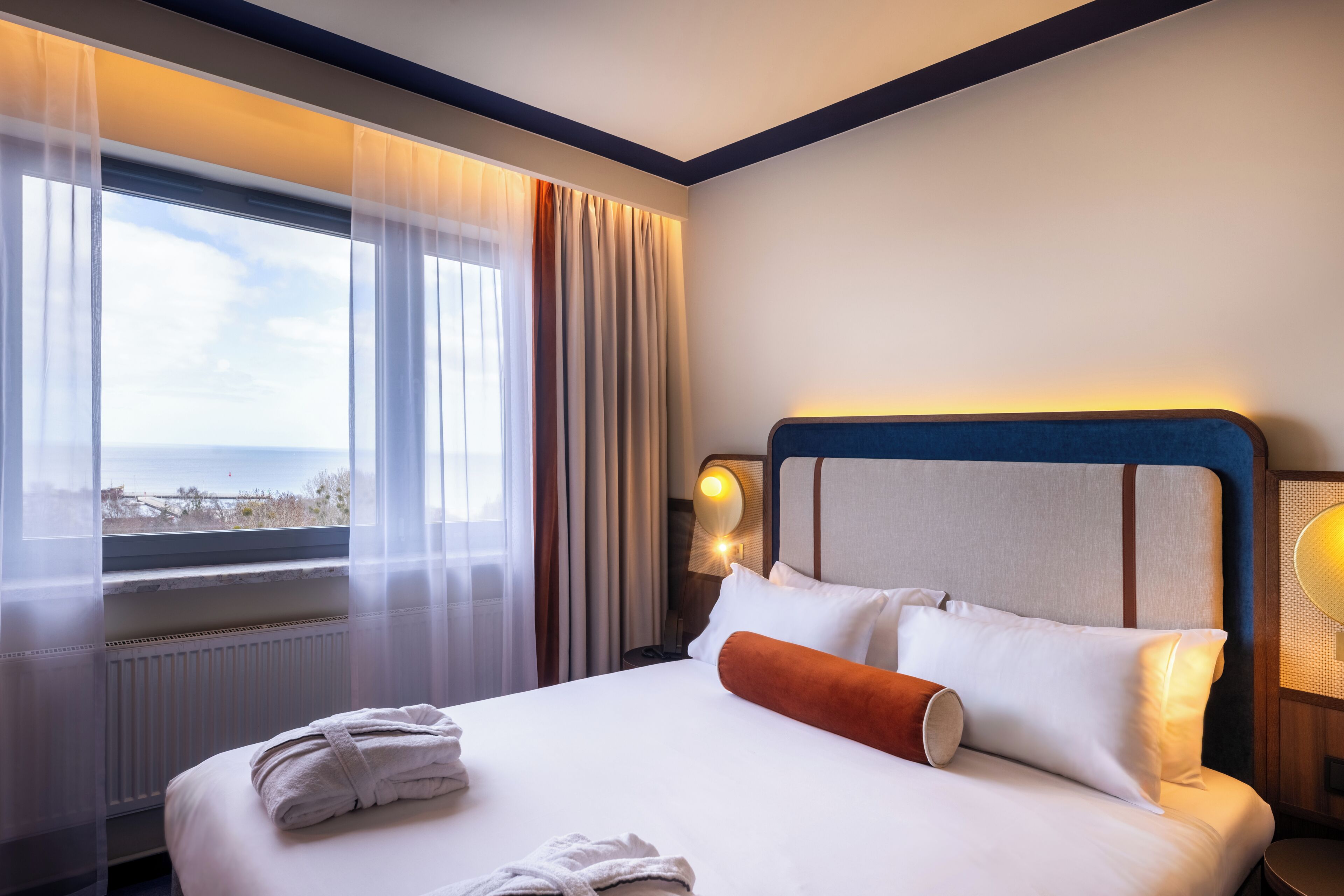 Een hotelkamer met een groot bed, uitzicht op zee, een raam met lichte gordijnen en een nachtkastje met een lamp.