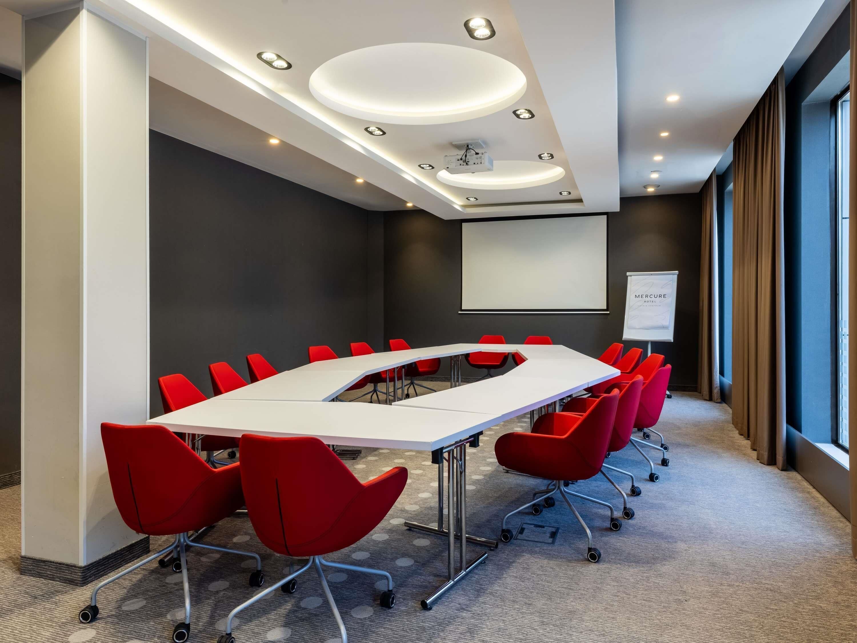 Une salle de conférence avec une longue table, des chaises rouges, un tableau blanc et un écran de projection.