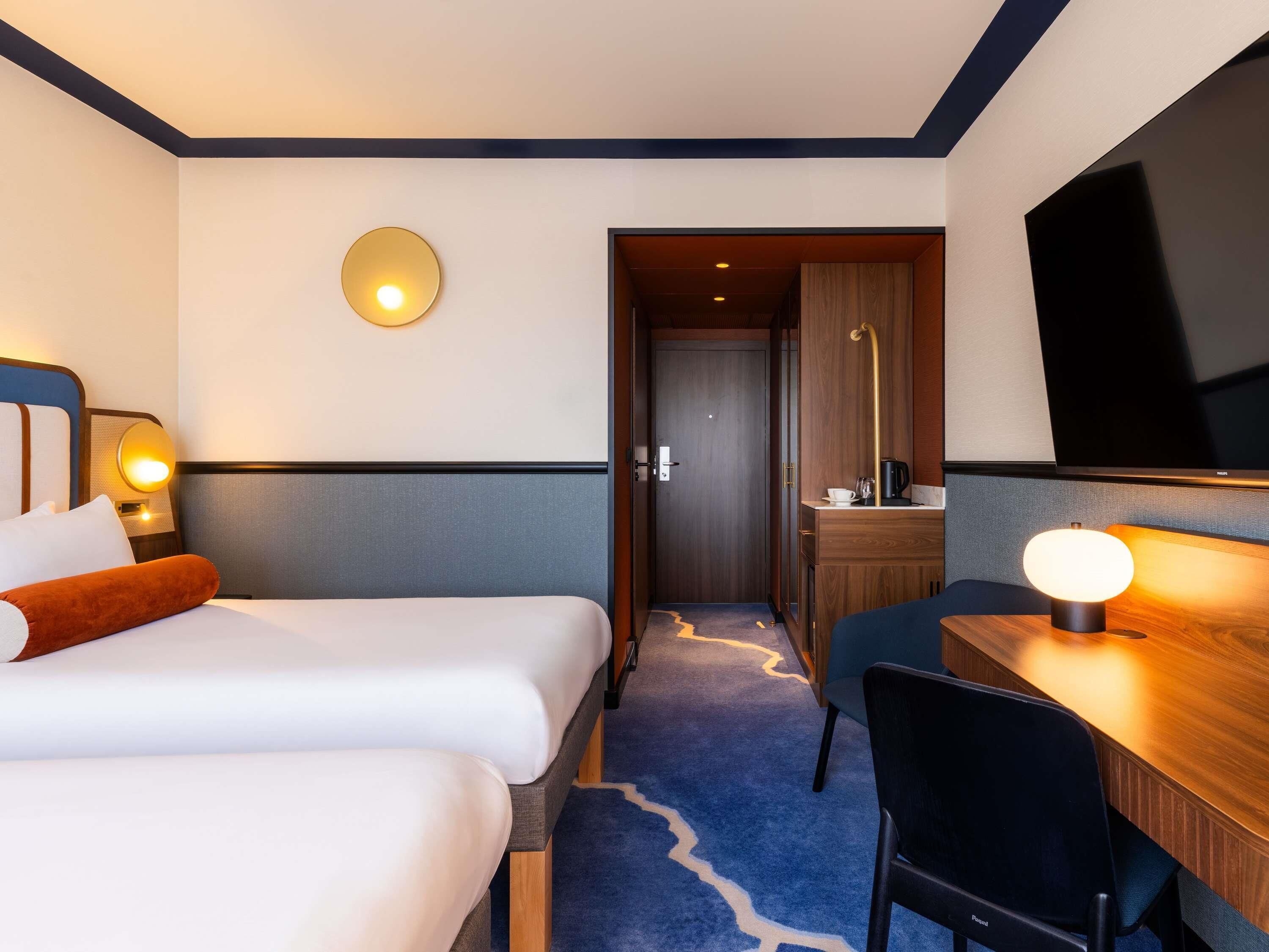 A hotel room with two beds, a desk, a chair, a TV, and a bathroom door.