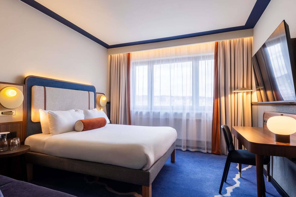A hotel room with a bed, bedside table, lamp, chair, desk, and a large window with curtains.