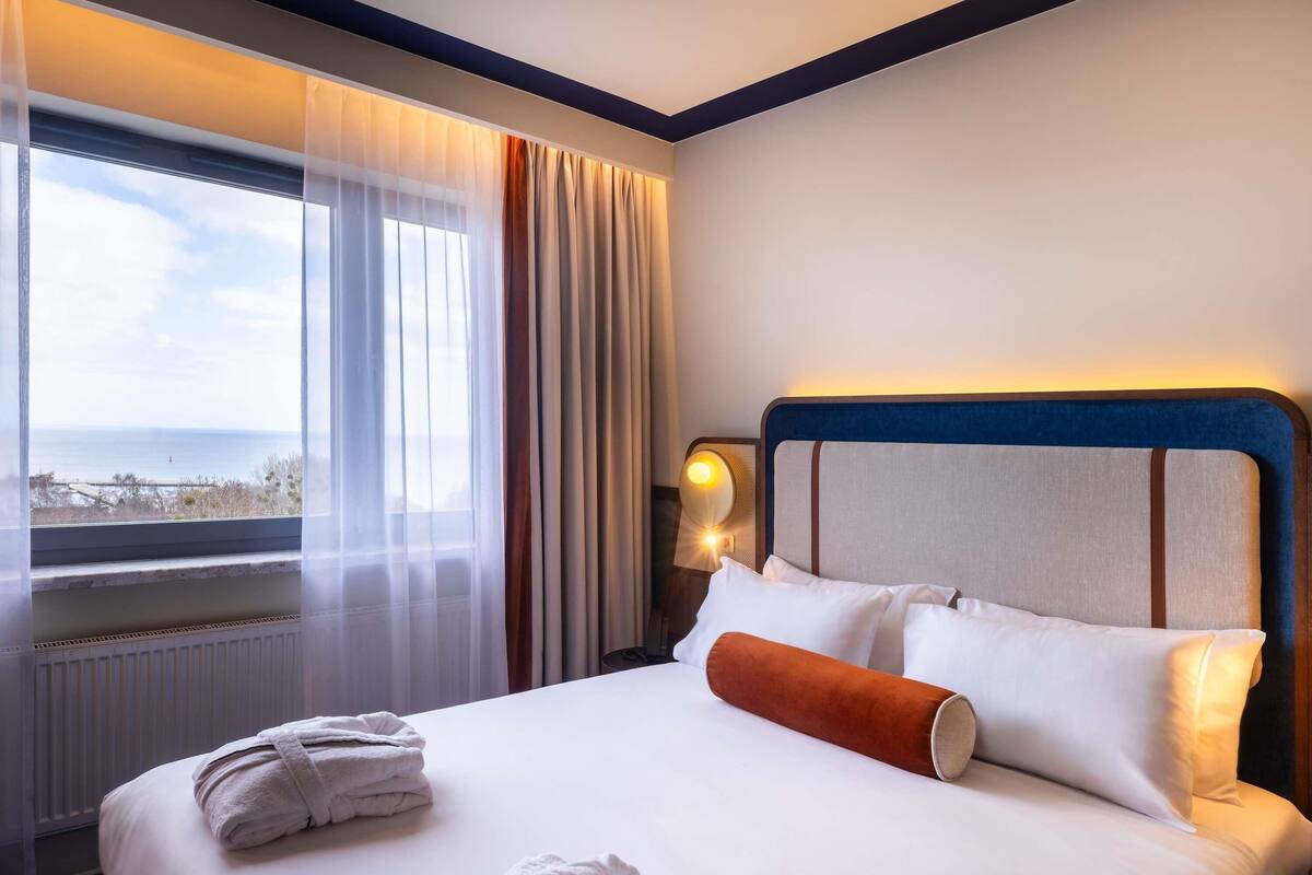 A hotel room with a bed, a nightstand, a lamp, a window with curtains, and a view of the sea.
