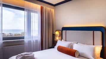 A hotel room with a bed, a nightstand, a lamp, a window with curtains, and a view of the sea.