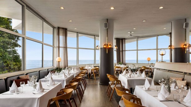 A restaurant with tables set for dining, large windows offering a view of the sea, and modern lighting fixtures.