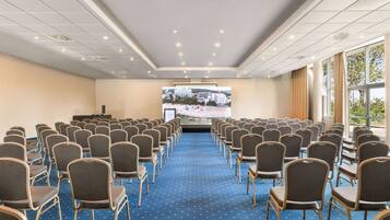 A conference room with rows of chairs facing a large screen, carpeted floor, and large windows.