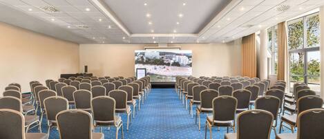 A conference room with rows of chairs facing a large screen, carpeted floor, and large windows.