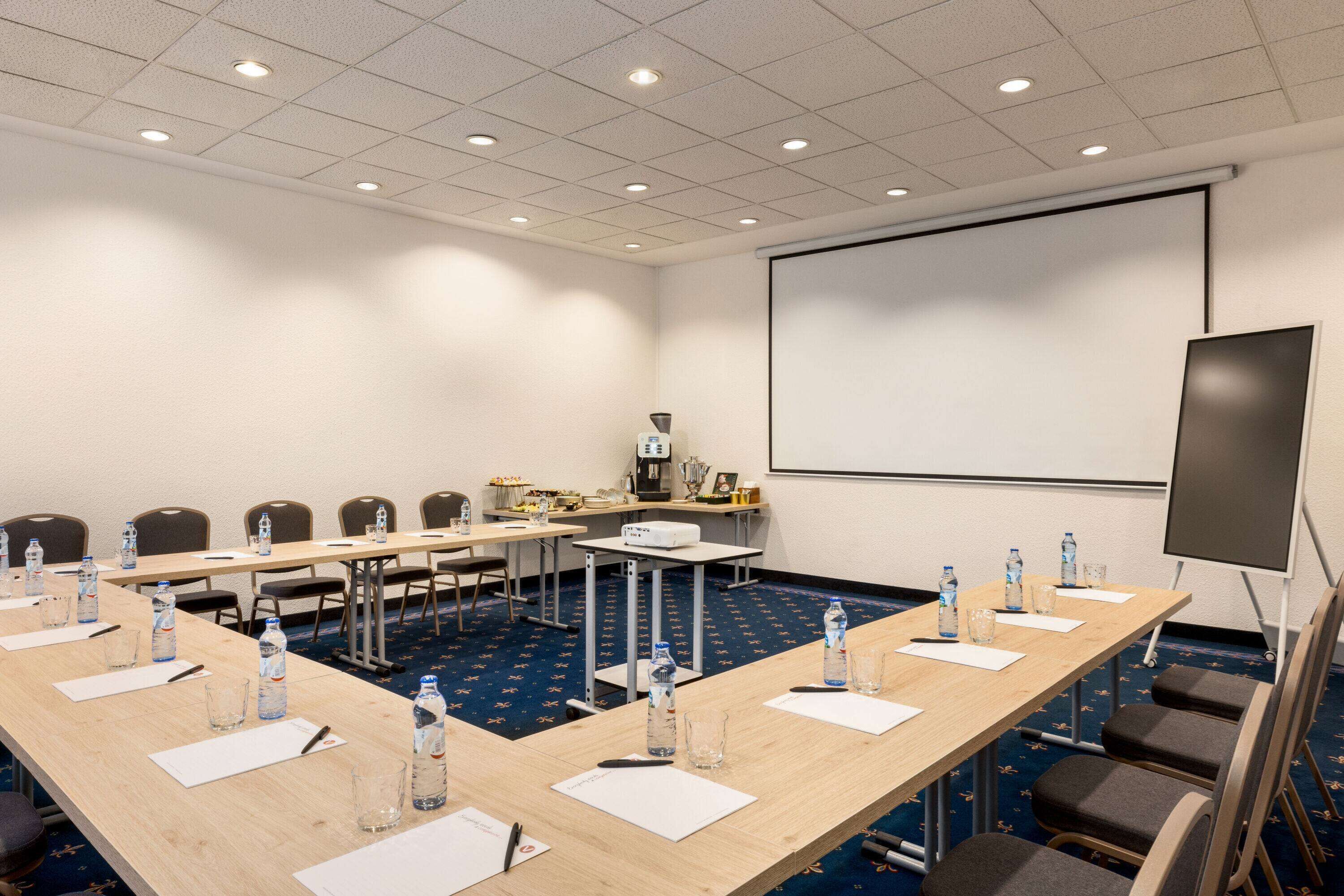 Un salón de conferencias con una larga mesa, sillas, botellas de agua y una pantalla de proyector.