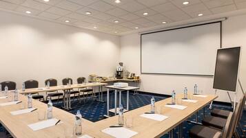 Un salón de conferencias con una larga mesa, sillas, botellas de agua y una pantalla de proyector.