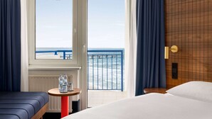 A hotel room with a bed, a small table with water bottles, a view of the ocean through a balcony door, and a wooden headboard.