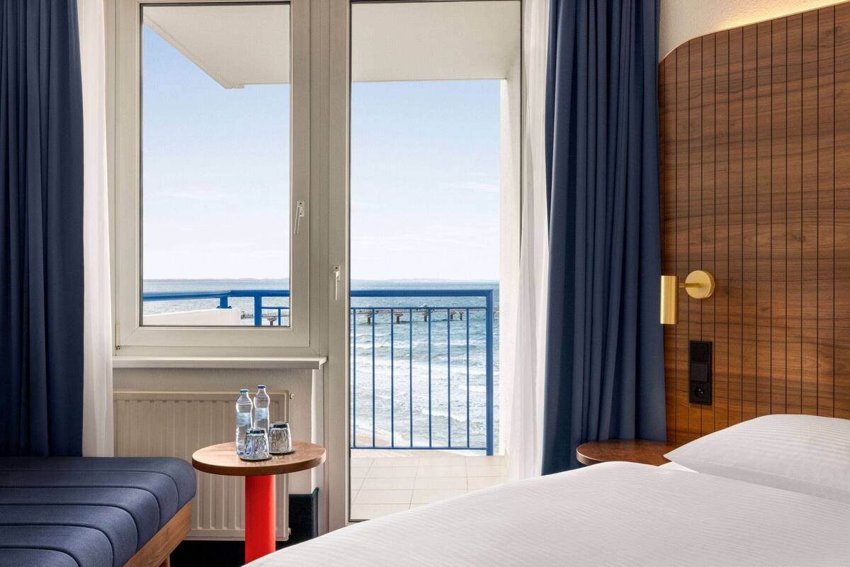Une chambre d’hôtel avec un lit, une petite table avec des bouteilles d’eau, une vue sur l’océan depuis la porte d’un balcon et une tête de lit en bois.