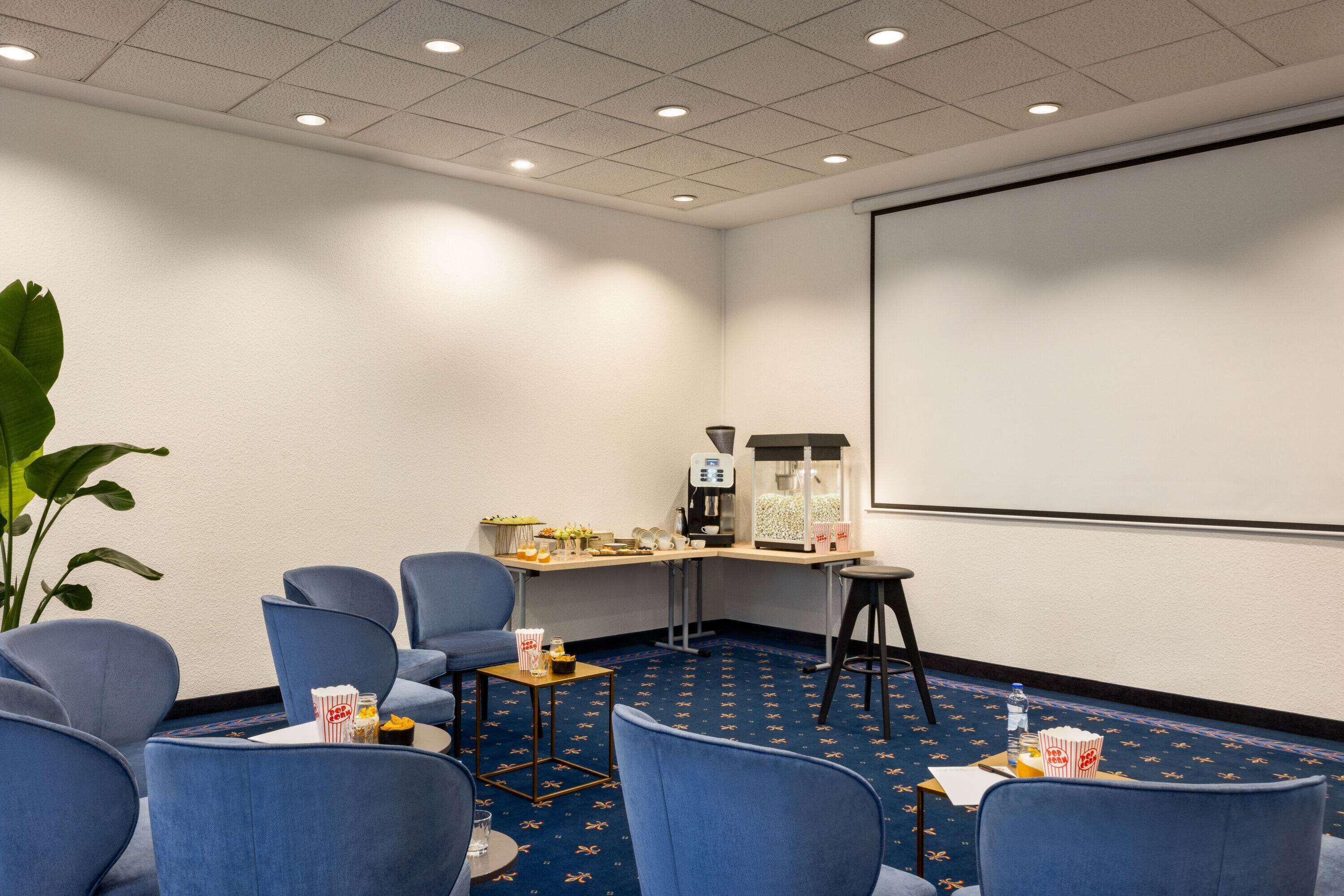A conference room with blue chairs, a projector screen, a coffee machine, and a small table with snacks.
