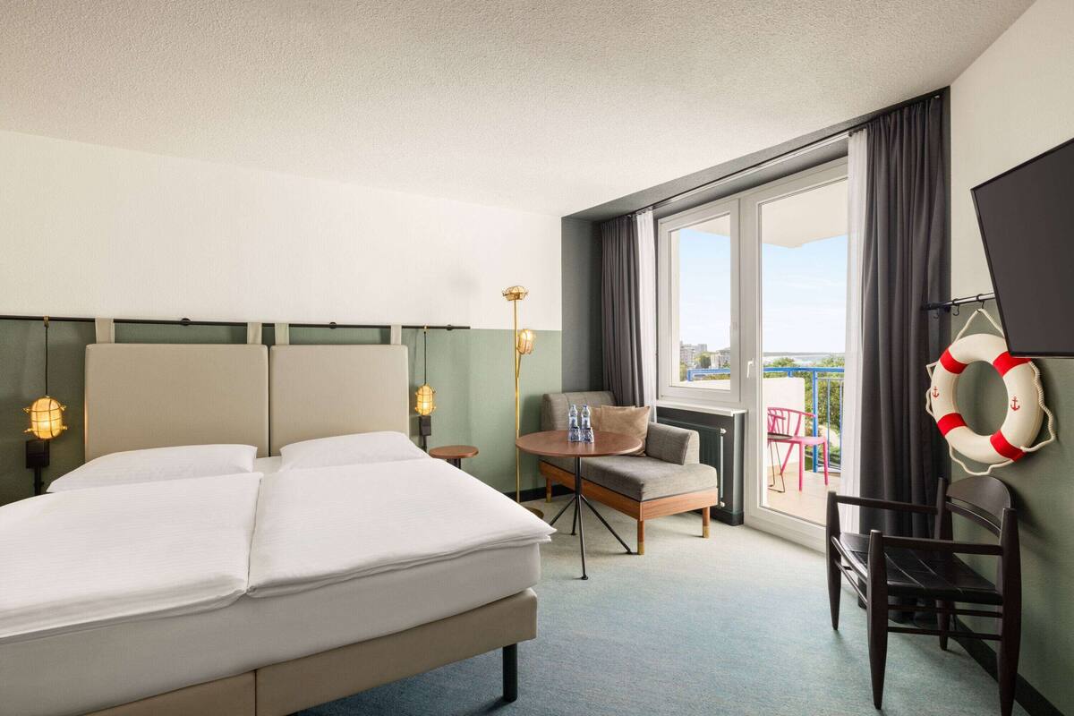Une chambre d’hôtel avec un lit, une petite table, une chaise, un canapé et une grande fenêtre donnant sur un paysage.