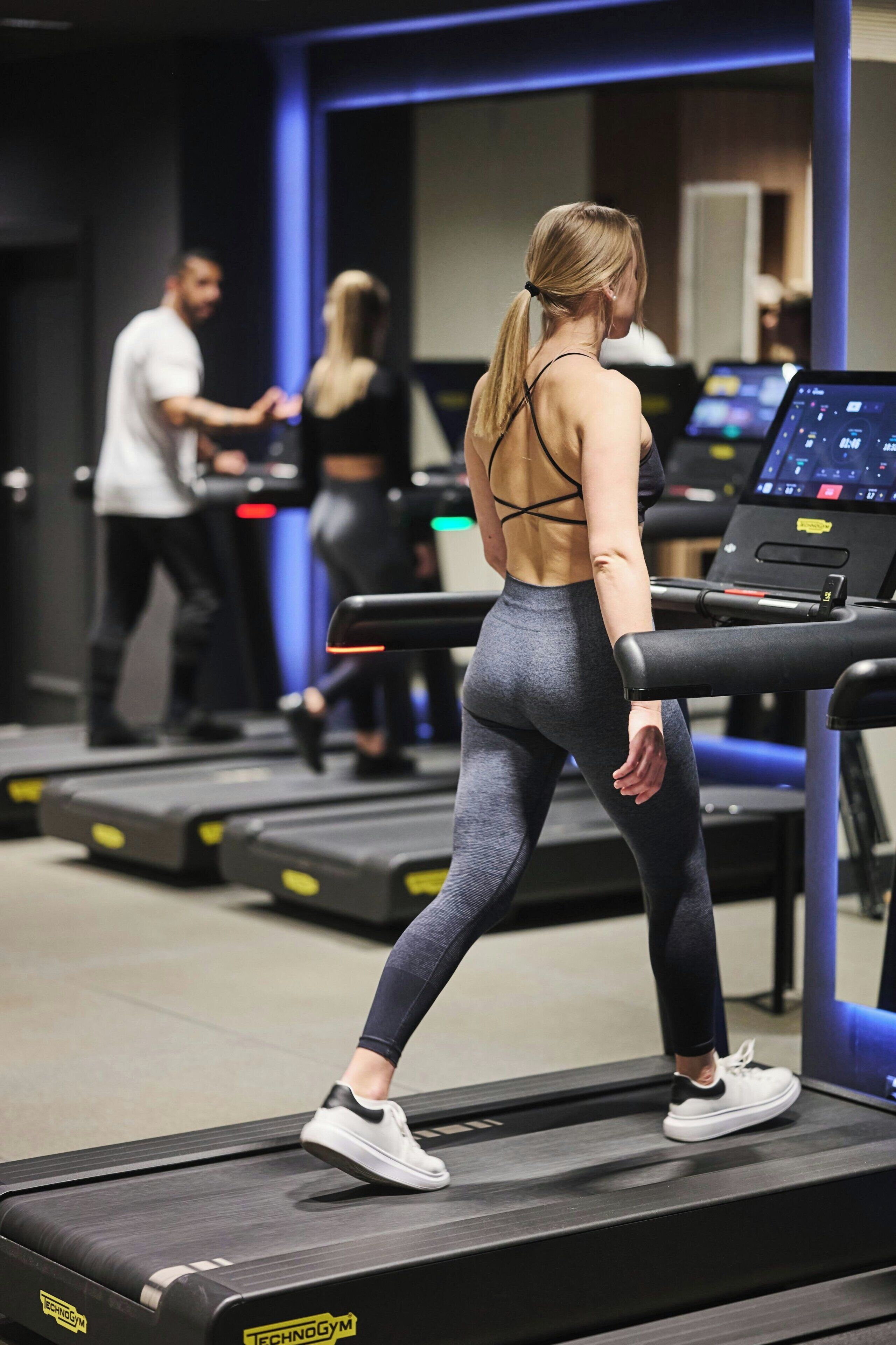 Une salle de sport avec des tapis de course, une femme qui marche sur l’un d’eux, et une autre personne à l’arrière-plan.