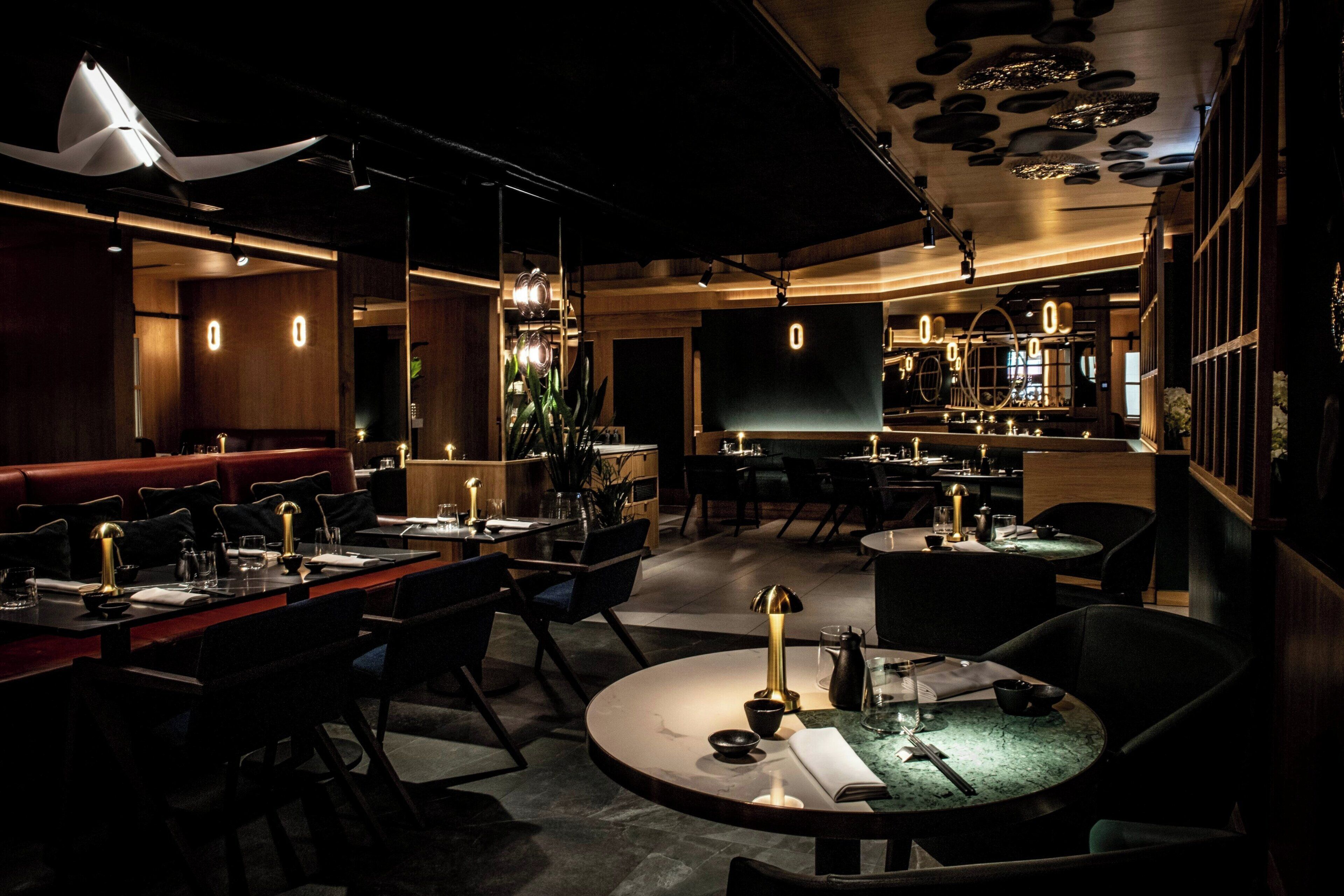 A dimly lit restaurant with round tables and booths, ambient lighting, and a modern interior design.