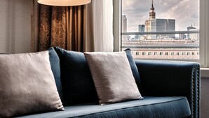 Una habitación de hotel moderna con un sofá azul oscuro, un ventanal con vistas a un horizonte urbano y un ventanal de techo a piso con cortinas.