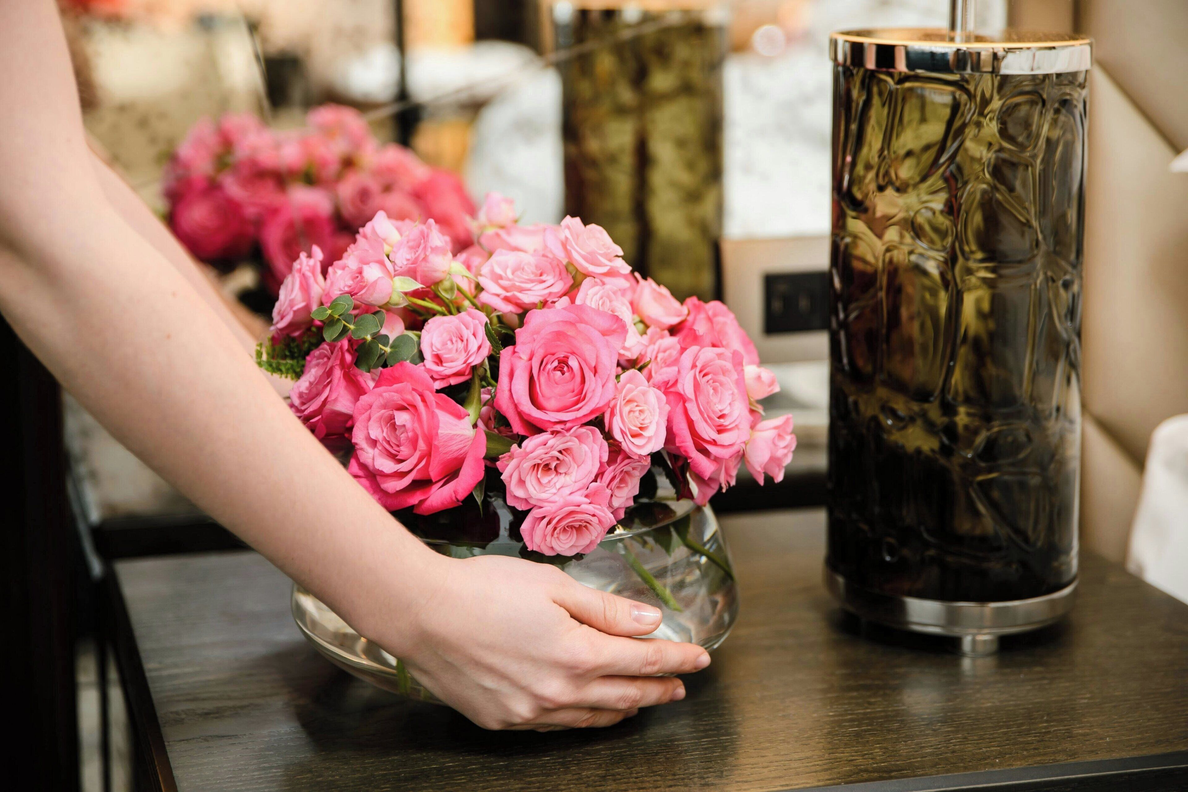 A hand holding a vase of pink roses on a wooden table next to a tall, cylindrical vase with a reflective surface.